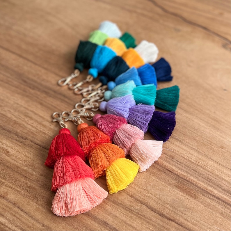May include: A set of colorful tassel keychains with gold metal rings. The tassels are arranged in a rainbow pattern, starting with light blue and ending with yellow.