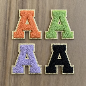 Peut inclure: Quatre écussons en forme de lettre A, de couleurs orange, vert lime, lavande et noir. Chaque écusson présente une surface texturée en chenille duveteuse avec une bordure à paillettes dorées, dans une police de caractères style varsity.