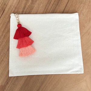 May include: A white canvas zippered pouch with a red, pink, and coral tassel attached to the zipper pull.
