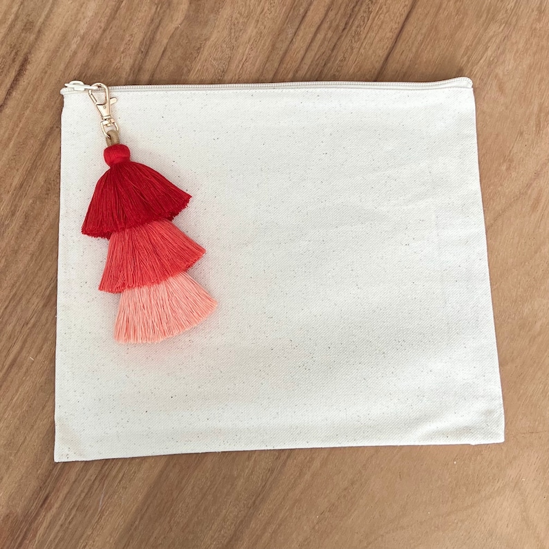 May include: A white canvas zippered pouch with a red, pink, and coral tassel attached to the zipper pull.