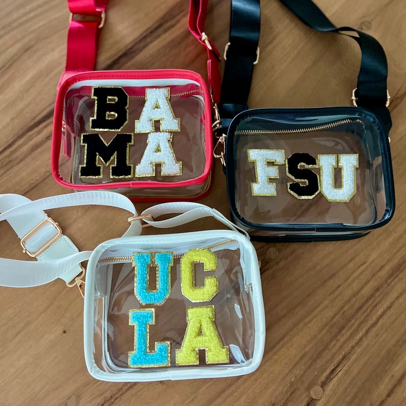 May include: Three clear plastic bags with different colored straps and gold zippers. Each bag has a different college team's name spelled out in colorful letters. The bags are on a wooden surface. The bags say "BAMA", "FSU", and "UCLA".