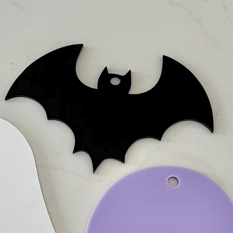 May include: A flat, black acrylic bat-shaped cutout with a small circular hole at the top for hanging. The bat features pointed ears and wide, scalloped wings. It is positioned above a partial white shape and a light purple circular piece.