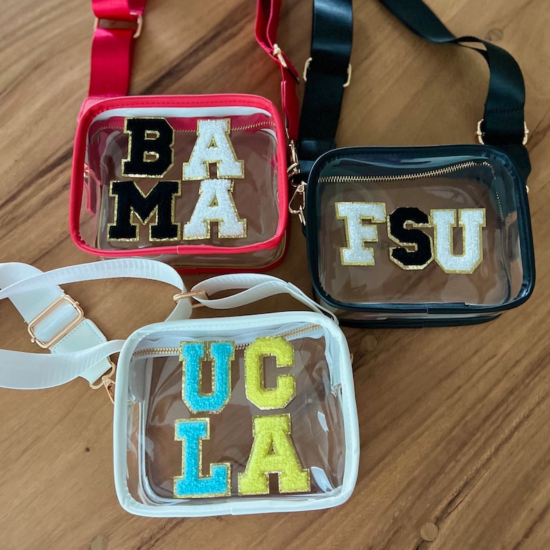 Op de afbeelding: Drie transparante plastic tassen met verstelbare banden. De tassen zijn versierd met kleurrijke vilt letters die "BAMA", "FSU" en "UCLA" spellen.