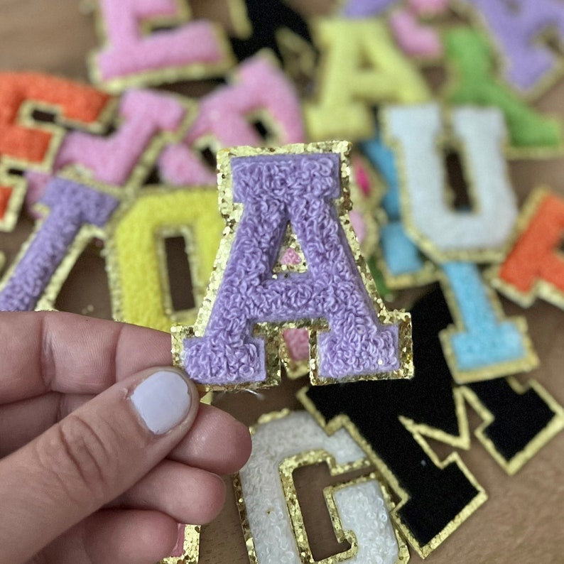 Op de afbeelding: Paarse viltletter A met gouden glitterrand. De letter is omringd door andere viltletters in verschillende kleuren met gouden glitterrand.