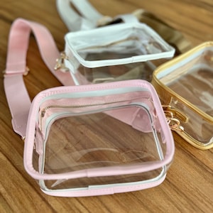 May include: Three clear plastic crossbody bags with different colored trim. The bags are pink, white, and gold. The bags have a zipper closure and a clear plastic body.