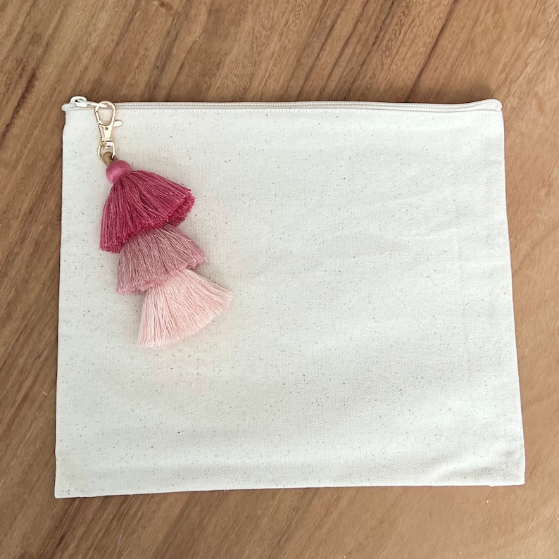 May include: A white canvas zippered pouch with a pink, ombre tassel attached to a gold key ring.
