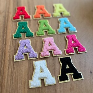 Op de afbeelding: Tien vilt letter patches in verschillende kleuren, elk met een gouden omtrek. De letters zijn allemaal de letter 'A' en zijn gerangschikt in twee rijen van vijf.