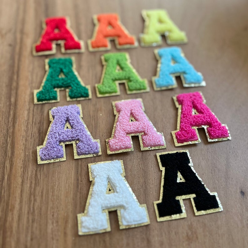 Op de afbeelding: Tien vilt letter patches in verschillende kleuren, elk met een gouden omtrek. De letters zijn allemaal de letter 'A' en zijn gerangschikt in twee rijen van vijf.