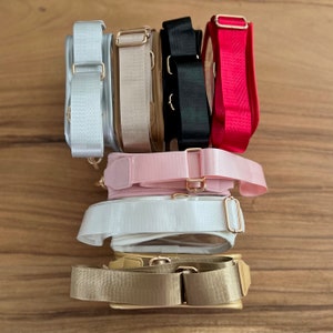May include: A stack of eight clear plastic bags with different colored straps. The straps are silver, beige, black, red, pink, white, and gold.