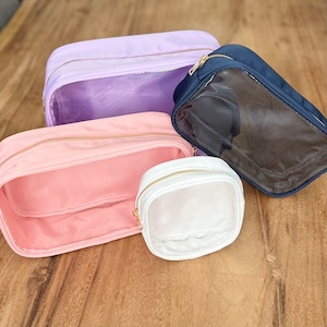 May include: Four clear makeup bags with zippers. The bags are in different sizes and colors: lavender, pink, white, and navy blue.