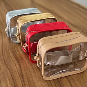 May include: Four clear crossbody bags with different colored leather trim. The bags are lined up in a row from left to right: light blue, gold, red, and beige.