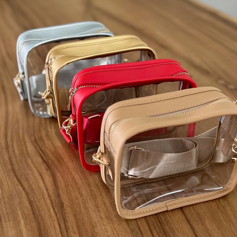 May include: Four clear crossbody bags with different colored leather trim. The bags are lined up in a row from left to right: light blue, gold, red, and beige.