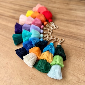 May include: A set of colorful tassel keychains with gold metal rings. The tassels are arranged in a rainbow pattern, starting with red and ending with white.
