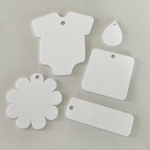 May include: A collection of five white, flat, smooth-surfaced shapes with pre-drilled holes for hanging. The set includes a babygrow shape, a teardrop, a square with rounded corners, a flower shape with multiple petals, and a rectangular tag.