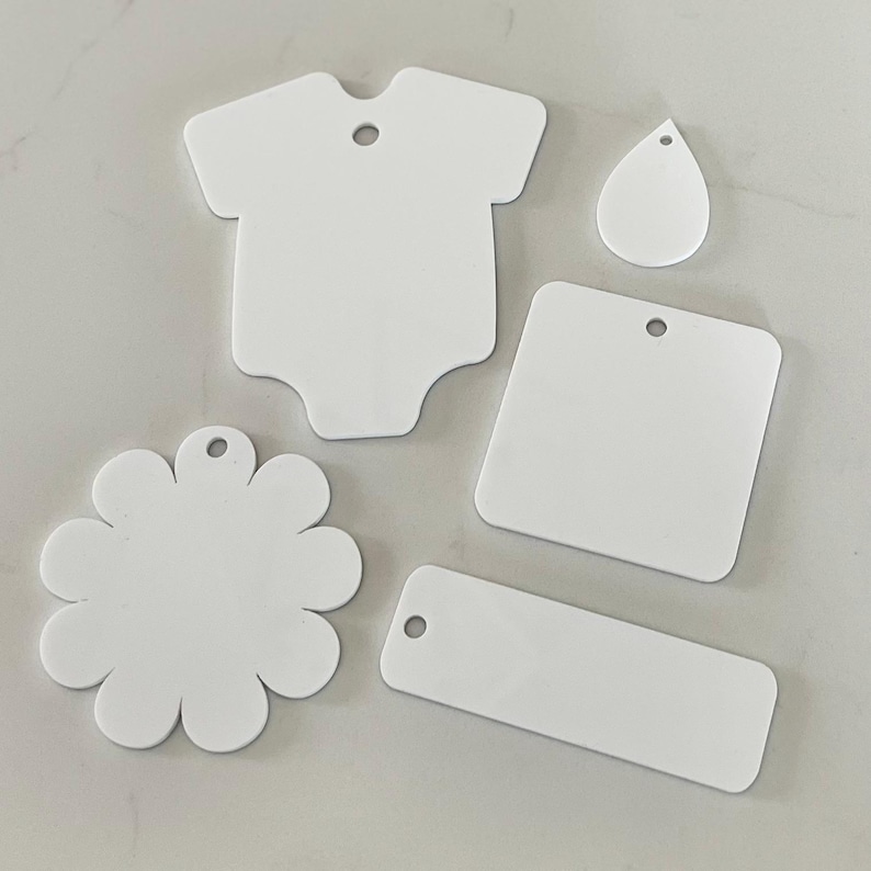 May include: A collection of five white, flat, smooth-surfaced shapes with pre-drilled holes for hanging. The set includes a babygrow shape, a teardrop, a square with rounded corners, a flower shape with multiple petals, and a rectangular tag.