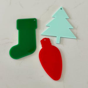 May include: Three flat acrylic ornaments featuring a dark green stocking, a light mint green Christmas tree, and a bright red light bulb shape. Each ornament has a small circular hole at the top for hanging, with smooth edges and a solid colour finish.