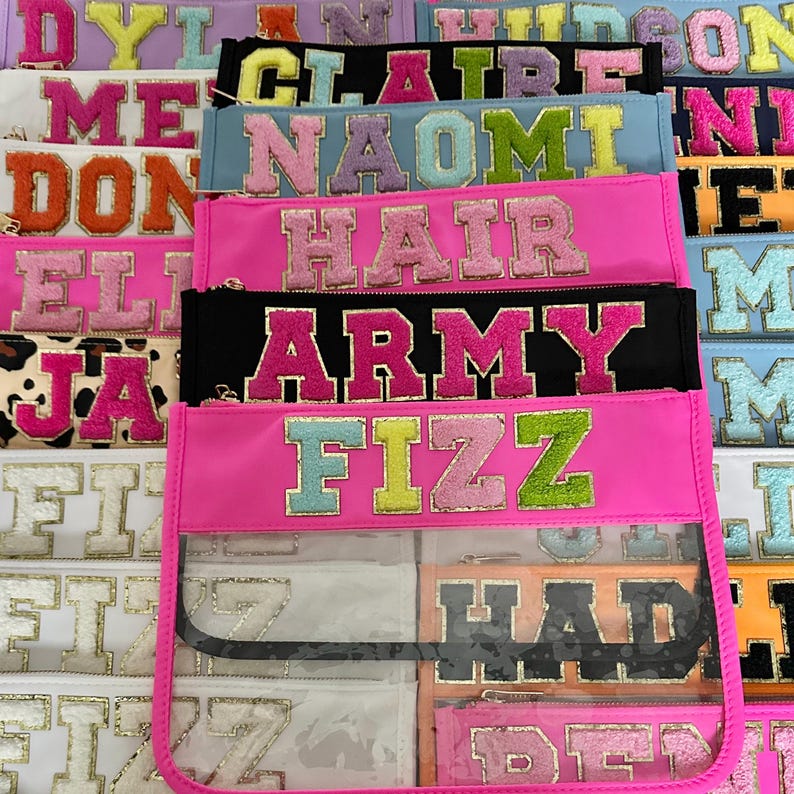 Pode incluir: Uma cole&ccedil;&atilde;o de bolsas com z&iacute;per coloridas com janelas de vinil transparente. Cada bolsa apresenta um nome diferente em letras coloridas e brilhantes. Os nomes incluem: Dylan, Claire, Naomi, Hair, Army, Fizz, Had e Ben.