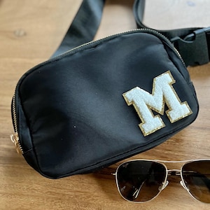 May include: Black nylon fanny pack with a gold zipper and a white and gold letter "M" patch. The bag has a black adjustable strap and a gold buckle. A pair of aviator sunglasses rests on the wooden surface.