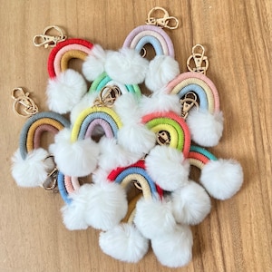 May include: A group of rainbow-colored keychain charms with fluffy white pom-poms. Each charm is made of yarn and features a rainbow arch with a gold key ring.
