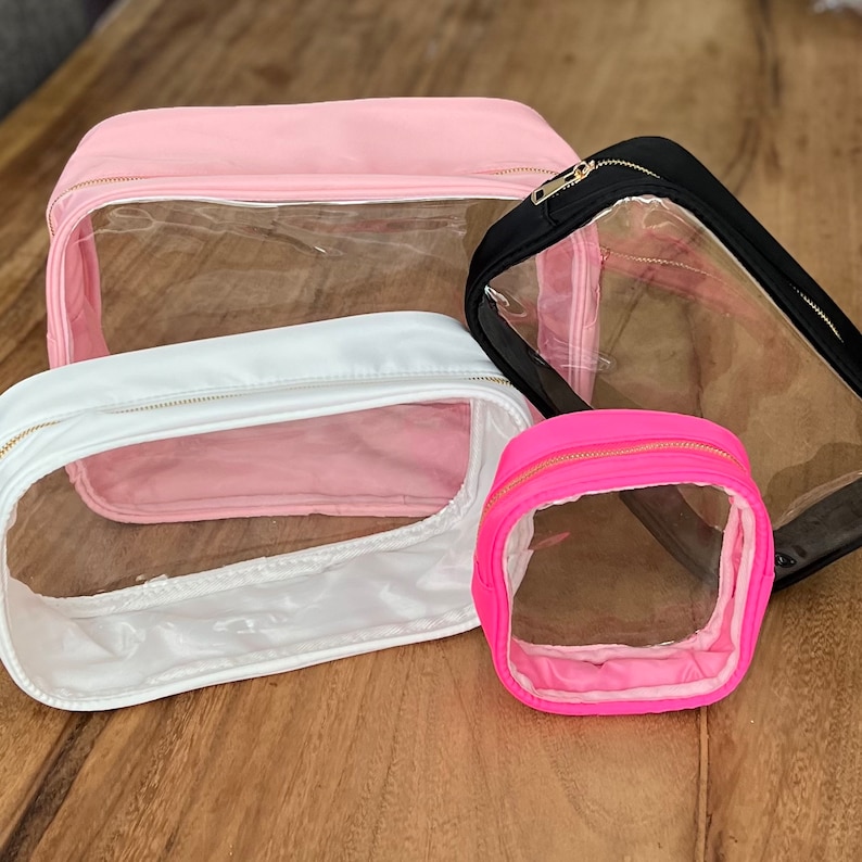 Op de afbeelding: Vier transparante ritszakjes met roze, witte en zwarte stoffen buitenkant. De zakjes zijn gemaakt van transparant vinyl en hebben gouden ritsen.