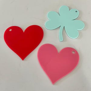 May include: Three flat acrylic shapes including one bright red heart, one light pink heart, and one mint green four-leaf clover. Each piece features a small circular hole near the top edge, suitable for hanging or attaching to other items.