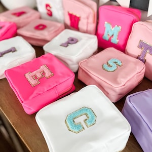 May include: A selection of small, square cosmetic bags in shades of pink, white, and lilac. Each bag is adorned with a letter applique in a contrasting colour, and has a gold zip. The bags are displayed on a wooden surface.