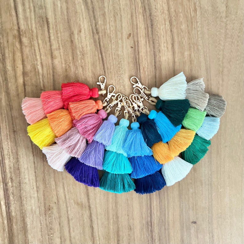 May include: A set of 15 colorful tassel keychains with gold key rings. The tassels are made of soft, fluffy yarn in a variety of colors, including red, pink, blue, green, and yellow.
