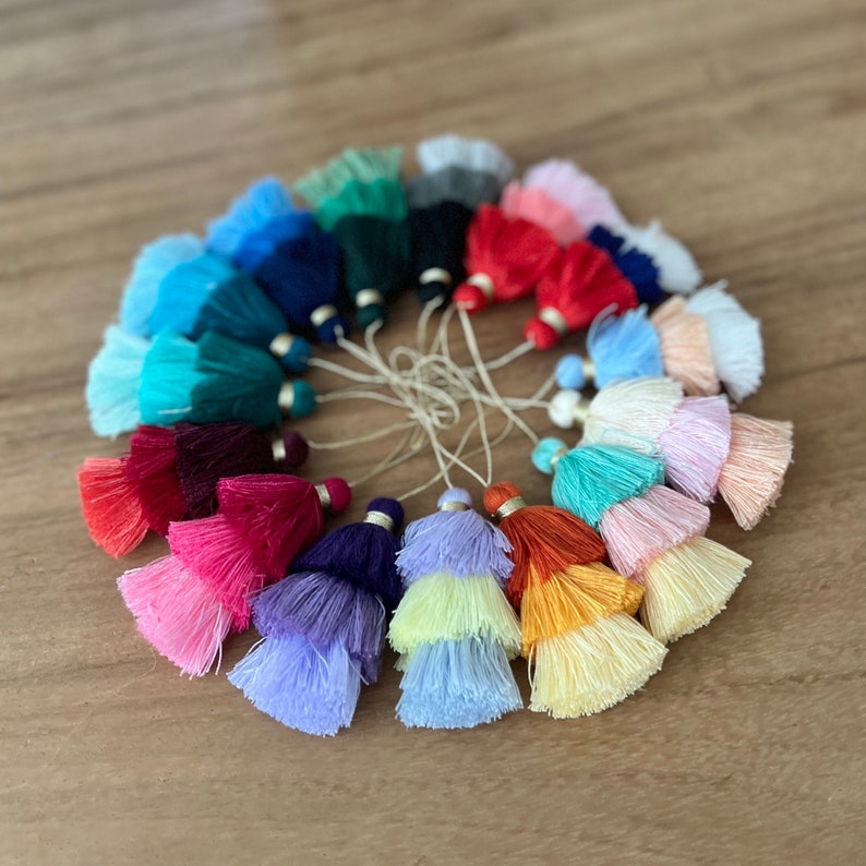 Pode incluir: Uma cole&ccedil;&atilde;o de 18 pingentes de franja coloridos em v&aacute;rios tons de azul, verde, vermelho, rosa, roxo e amarelo. As franjas est&atilde;o dispostas em c&iacute;rculo sobre uma superf&iacute;cie de madeira.