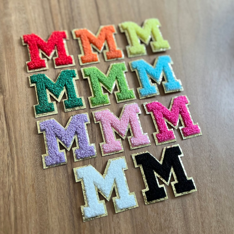 Op de afbeelding: Een set van tien vilt letter patches in verschillende kleuren, elk met de letter "M" in een groot, vet lettertype. De patches zijn in een verspringend patroon gerangschikt, waarbij de kleuren overgaan van rood naar groen naar blauw naar roze naar wit naar zwart.