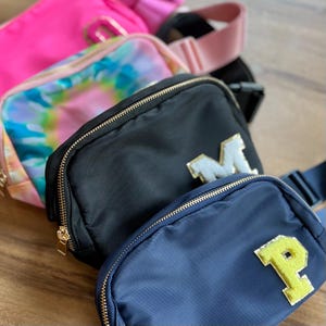 May include: Four different fabric fanny packs with gold zippers. The fanny packs are in pink, tie-dye, black, and navy blue. Each fanny pack has a large letter on the front, M, P, and two others.