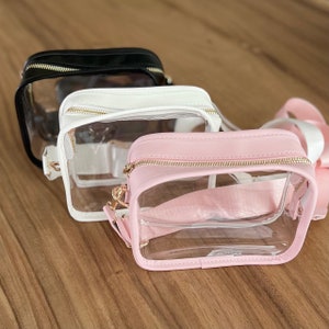 May include: Three clear plastic crossbody bags with gold zippers and straps. The bags are in black, white, and pink.