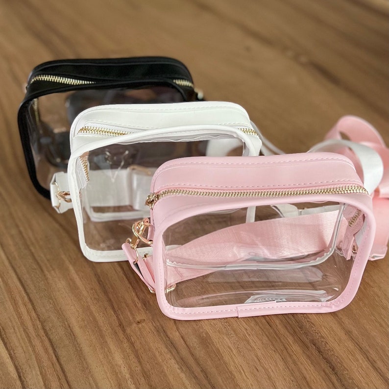 May include: Three clear plastic crossbody bags with gold zippers and straps. The bags are in black, white, and pink.
