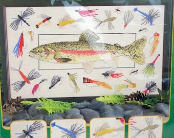 May include: A Nature's Window cross-stitch kit featuring a central rainbow trout surrounded by various colourful fishing flies. The packaging displays the finished design on a cream-coloured fabric background, with additional close-up images of individual fly patterns at the bottom and top right corner.