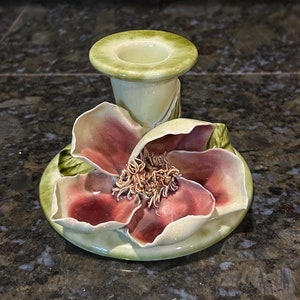 May include: A ceramic candle holder featuring a detailed magnolia flower design. The flower has burgundy petals and a textured center, set on a green base with a matching candle cup. The piece is hand-painted with a glossy finish.