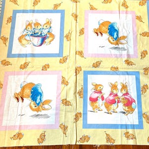 May include: A yellow fabric featuring four square panels with illustrations of rabbits in various poses. The panels are framed in blue and pink, with a repeating pattern of small rabbit figures scattered across the fabric.