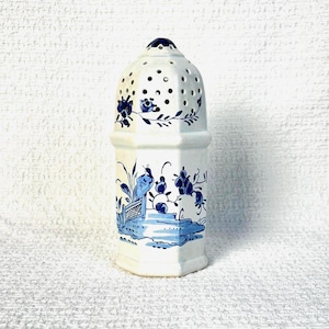 May include: A white ceramic shaker featuring a hexagonal body and a domed top with perforated holes. The surface is decorated with blue painted illustrations of birds, floral motifs, and landscape elements, including a small fence and foliage.