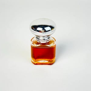 May include: A small, square glass perfume bottle filled with a dark amber liquid. It has a silver, rounded cap. The bottle is set against a plain white background, highlighting its shape and color.