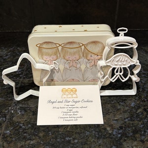 May include: A white metal tin with a picture of three angels on the front. The tin contains three cookie cutters: a star, an angel, and a bell. A recipe card for angel and star sugar cookies is included. The recipe calls for 1 cup sugar, 3/4 cup butter or margarine, 2 eggs, 1 teaspoon vanilla, 2 1/2 cups flour, 1 teaspoon baking powder, and 1 teaspoon salt.