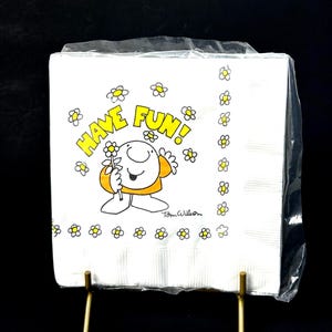 May include: A stack of white paper napkins with a cartoon illustration. The illustration features a smiling character holding a flower, surrounded by daisies, and the words "HAVE FUN!" in yellow. The napkins are in a clear plastic wrapper.