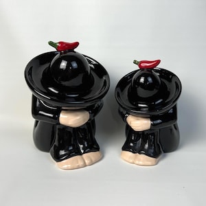 May include: Two ceramic figurines of a mariachi musician, one large and one small. The figures are dressed in black outfits with wide-brimmed hats. Each hat is topped with a red chili pepper. The figures have tan hands and feet.
