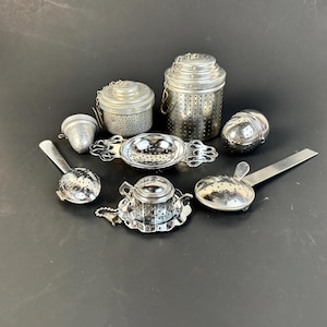 May include: A collection of seven silver-toned metal tea infusers and strainers in various shapes, including cylindrical containers, a small teapot-shaped infuser, a spherical ball, and handled strainers, all featuring perforated surfaces.