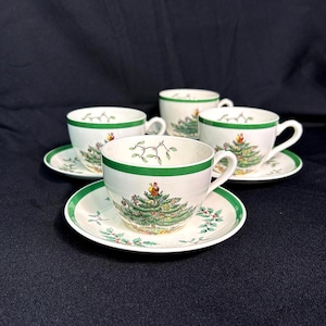 May include: Four white teacups and saucers with a green and white Christmas tree design. The cups and saucers have a green border. The teacups are on a black surface.