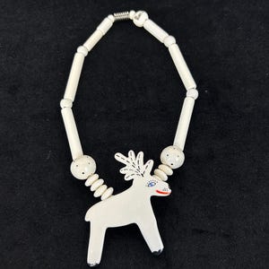 May include: A necklace featuring a white ceramic reindeer pendant with black antlers, a blue eye, and a red mouth. The chain consists of long white cylindrical beads, small white round beads with black dots, and flat white oval beads on a metal wire.
