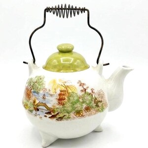 May include: White ceramic teapot with a green lid and a metal handle. The teapot features a scenic landscape design with a bridge, trees, and buildings. The teapot has three small feet and a spout.