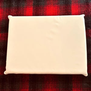 Pu&ograve; includere: Un cuscino rettangolare bianco sporco con angoli arrotondati. Il cuscino &egrave; posto su un tessuto a fantasia scozzese rosso e nero. La superficie del cuscino appare liscia e senza decorazioni, offrendo un'estetica pulita e minimalista.
