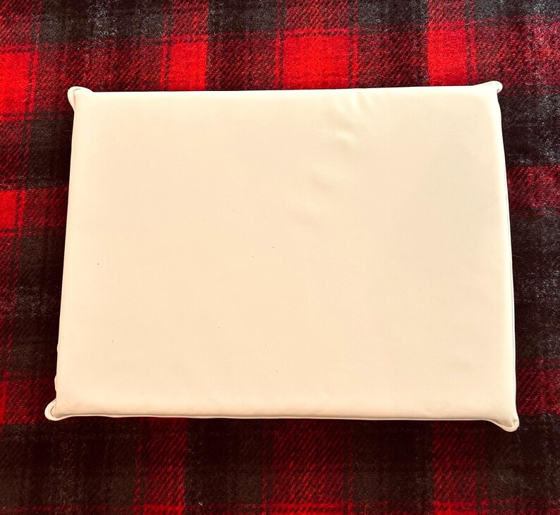 Pu&ograve; includere: Un cuscino rettangolare bianco sporco con angoli arrotondati. Il cuscino &egrave; posto su un tessuto a fantasia scozzese rosso e nero. La superficie del cuscino appare liscia e senza decorazioni, offrendo un'estetica pulita e minimalista.