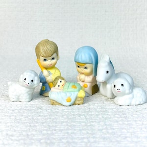 May include: A ceramic nativity figurine set featuring a central infant in a manger, two figures with painted facial features, a white sheep, and a white donkey. The figures are decorated with soft pastel colors including blue, yellow, and green accents.