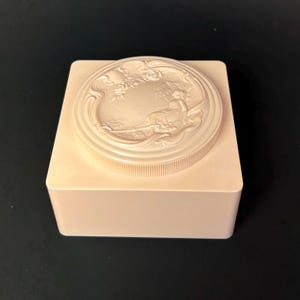 May include: A light pink, square-shaped trinket box with a circular lid. The lid features a detailed relief design of a seated figure in a landscape. The box has a ribbed edge on the lid and a smooth, matte finish.