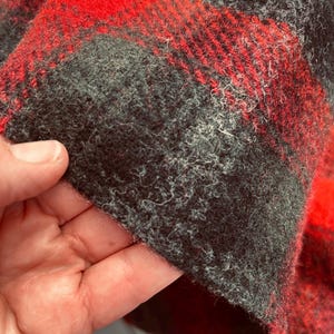 Pu&ograve; includere: Primo piano di un tessuto a quadri rosso e nero. Il tessuto ha un aspetto strutturato e sfocato. Il motivo &egrave; composto da audaci quadrati e linee rosse e nere. Il tessuto &egrave; probabilmente lana o un materiale simile.