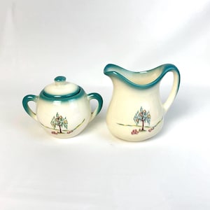 May include: A cream-colored ceramic sugar bowl with a lid and a matching creamer pitcher. Both pieces feature a hand-painted willow tree design with yellow accents and a green gradient rim. The sugar bowl has two side handles and a small lid knob.